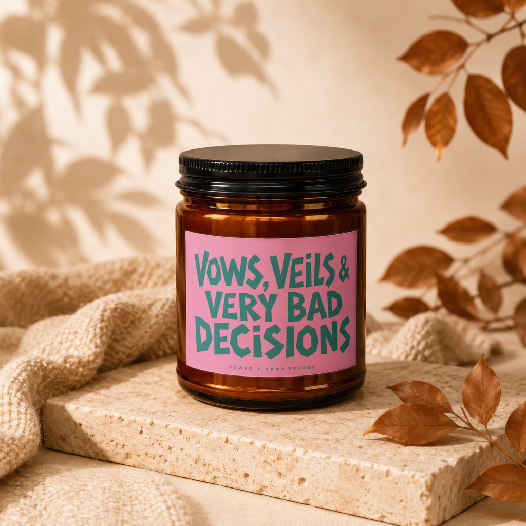 Amber jar candle with pink label reading "VOWS, VEILS & VERY BAD DECISIONS".