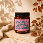 Amber jar candle with pink label reading "VOWS, VEILS & VERY BAD DECISIONS".