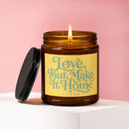 Amber glass candle with lit wick and black lid, features "Love, But Make It Home" text.
