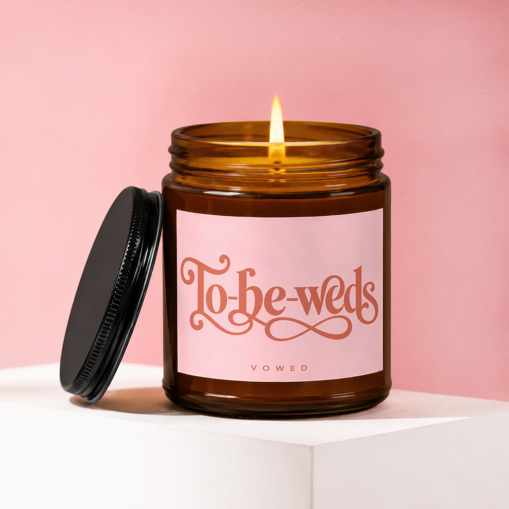 Lit amber glass candle with "To-be-weds" and "vowed" on pink label.