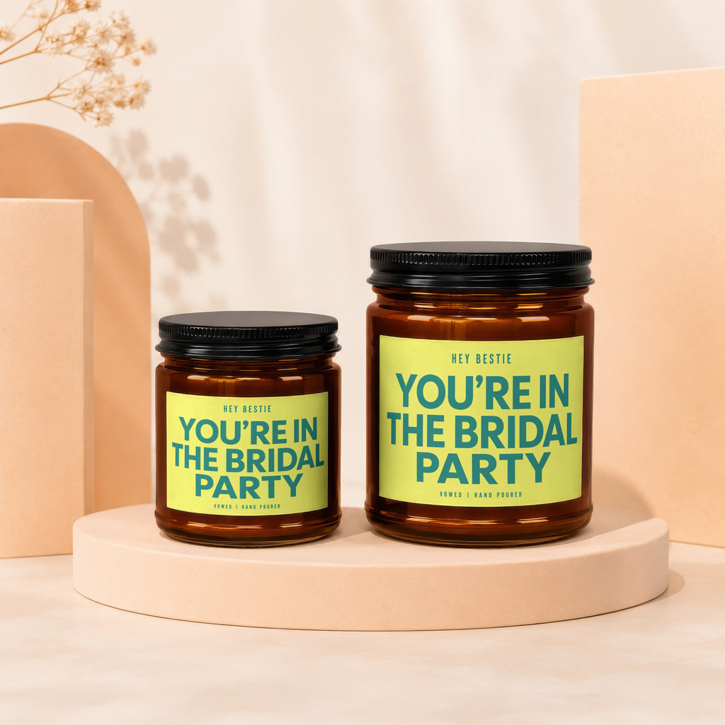 Two amber jars with black lids, featuring bright yellow labels that read "HEY BESTIE YOU'RE IN THE BRIDAL PARTY."