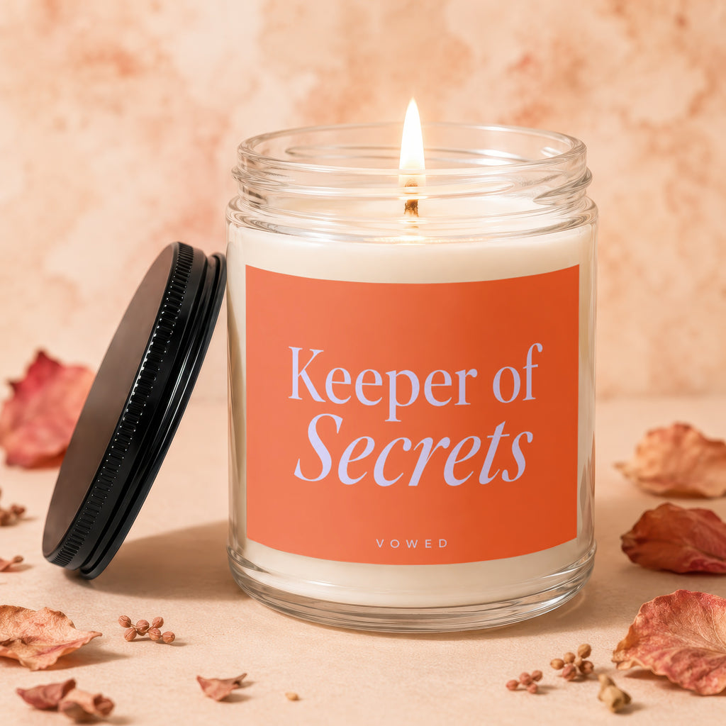 Glass candle with lit flame and orange label reading "Keeper of Secrets"