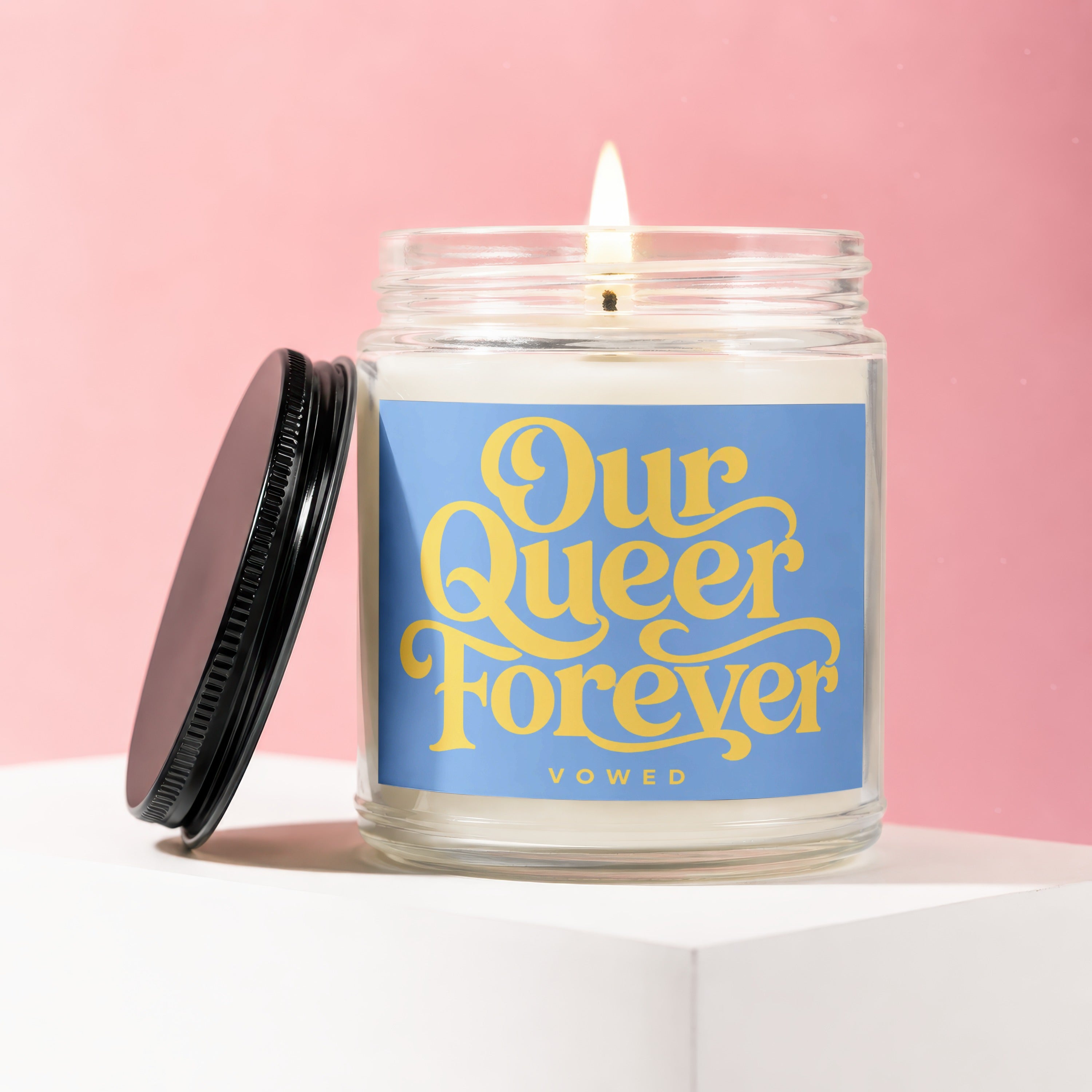 Glass candle with lit wick, blue label reads "Our Queer Forever Vowed".