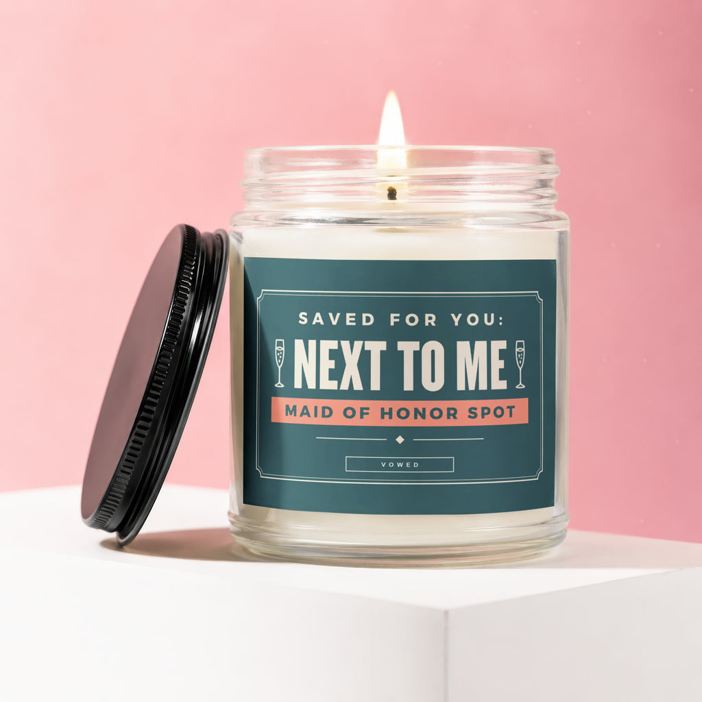 Candle labeled "NEXT TO ME MAID OF HONOR SPOT", lid off, on white surface.