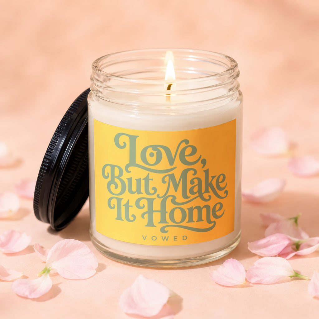 Glass candle with lit wick and "Love, But Make It Home" text on label.