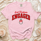 Light pink t-shirt featuring the text First Christmas Engaged above a diamond ring graphic, displayed on a textured cloth surface.