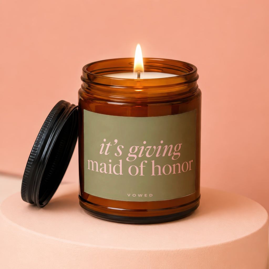 Amber candle with lit wick and black lid, reads 'it's giving maid of honor'.