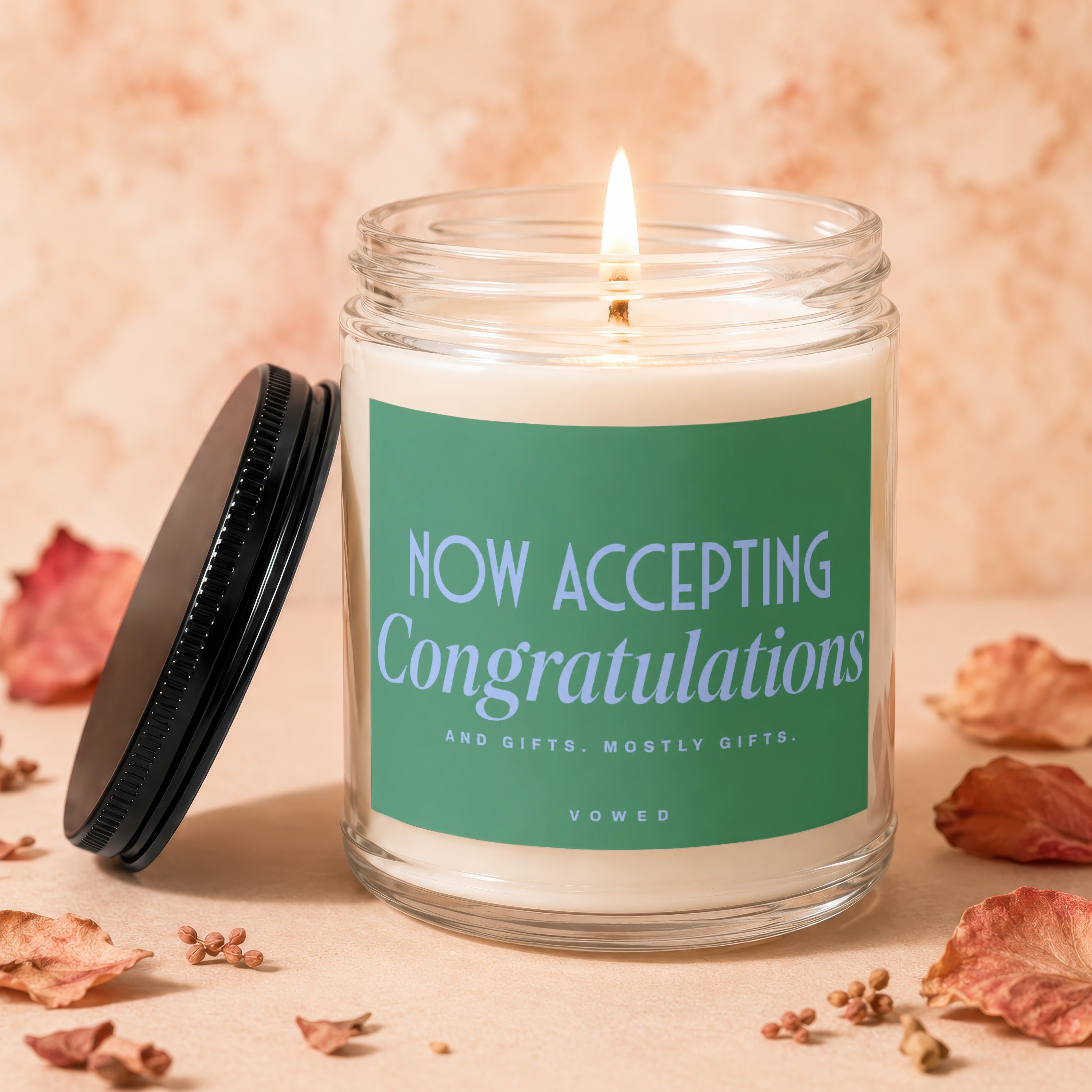 Lit candle in glass jar with "NOW ACCEPTING Congratulations and gifts. mostly gifts. VOWED" on label.