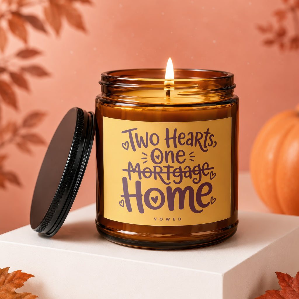Amber jar candle with lit wick and label reading "Two Hearts, One Mortgage, Home".