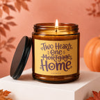Amber jar candle with lit wick and label reading "Two Hearts, One Mortgage, Home".