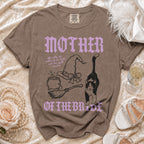 Brown crewneck t-shirt with purple and black graphics, witch-themed text, and accessories.