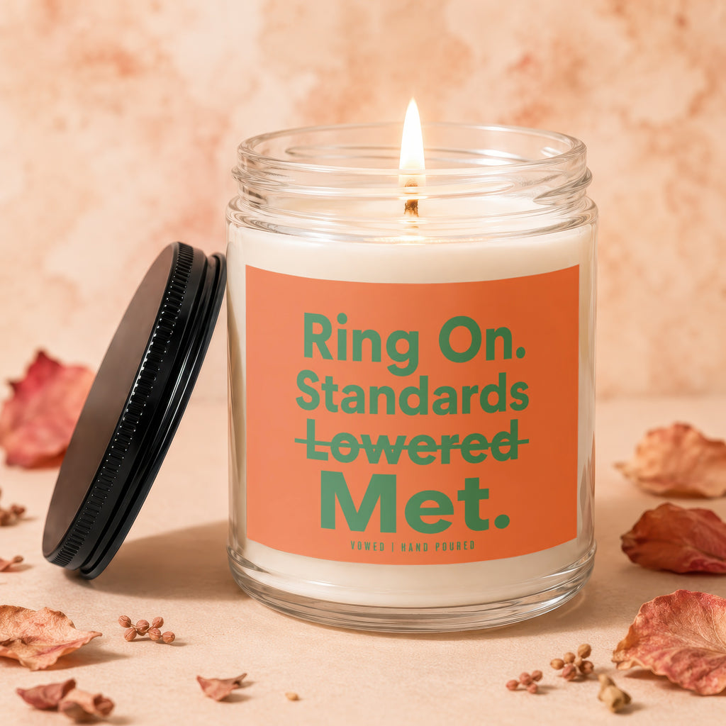 Glass candle with lit wick and orange label that reads "Ring On. Standards Lowered Met."