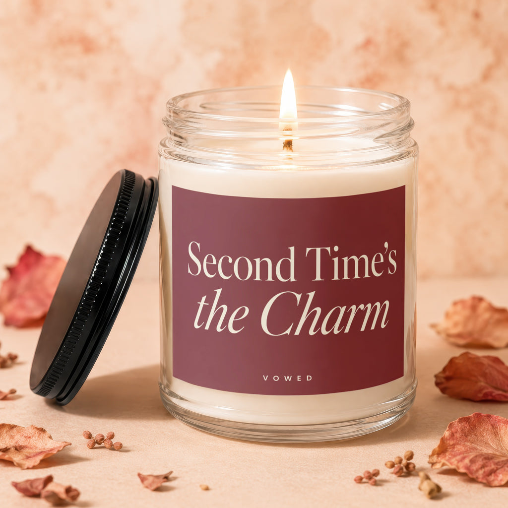 Glass candle with lit wick and label reading "Second Time's the Charm"