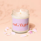 Lit candle in glass jar with "enGAYged" and "vowed" on label, surrounded by pink petals.
