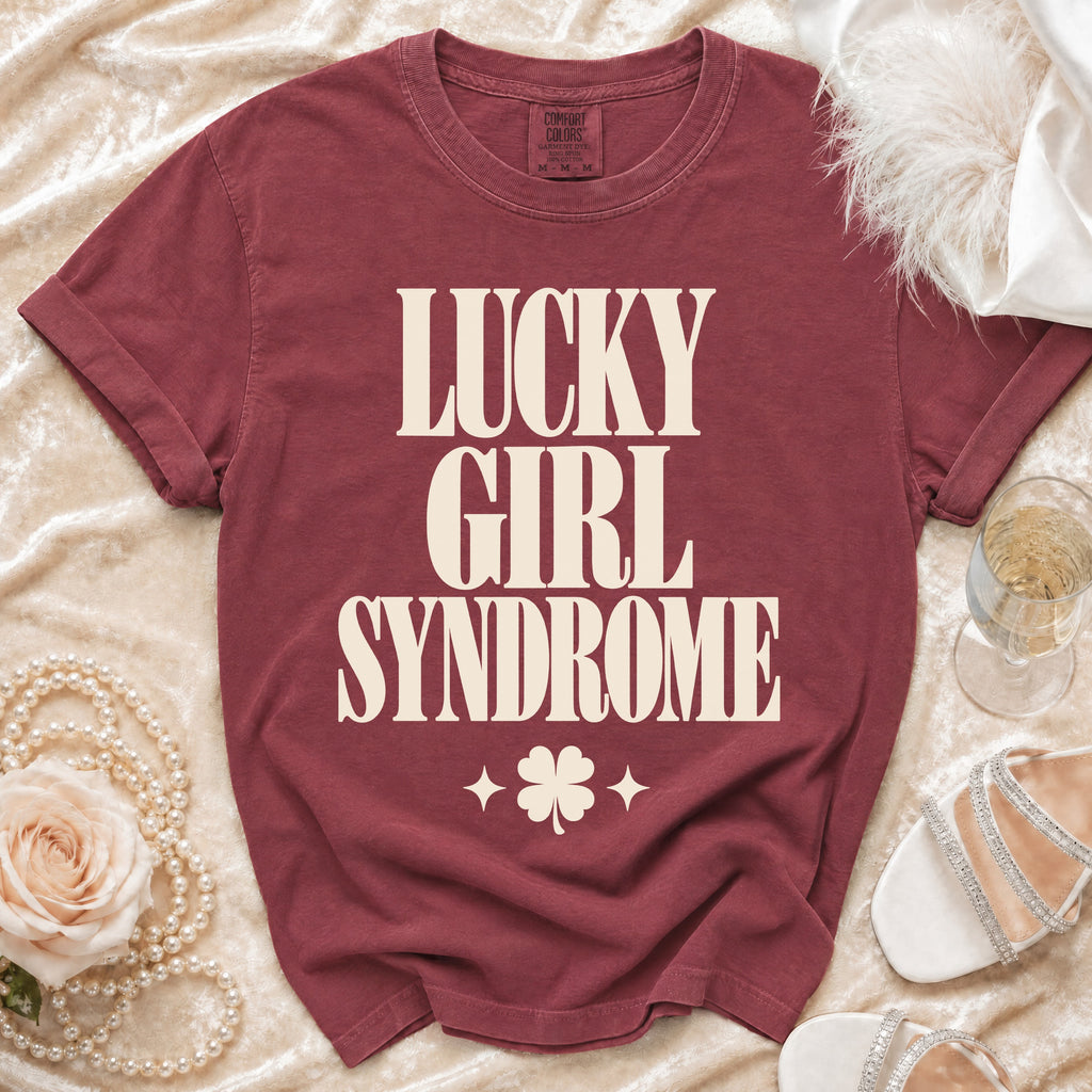 Maroon t-shirt with cream text reading "LUCKY GIRL SYNDROME" and a four-leaf clover, laid flat.
