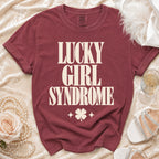 Maroon t-shirt with cream text reading "LUCKY GIRL SYNDROME" and a four-leaf clover, laid flat.