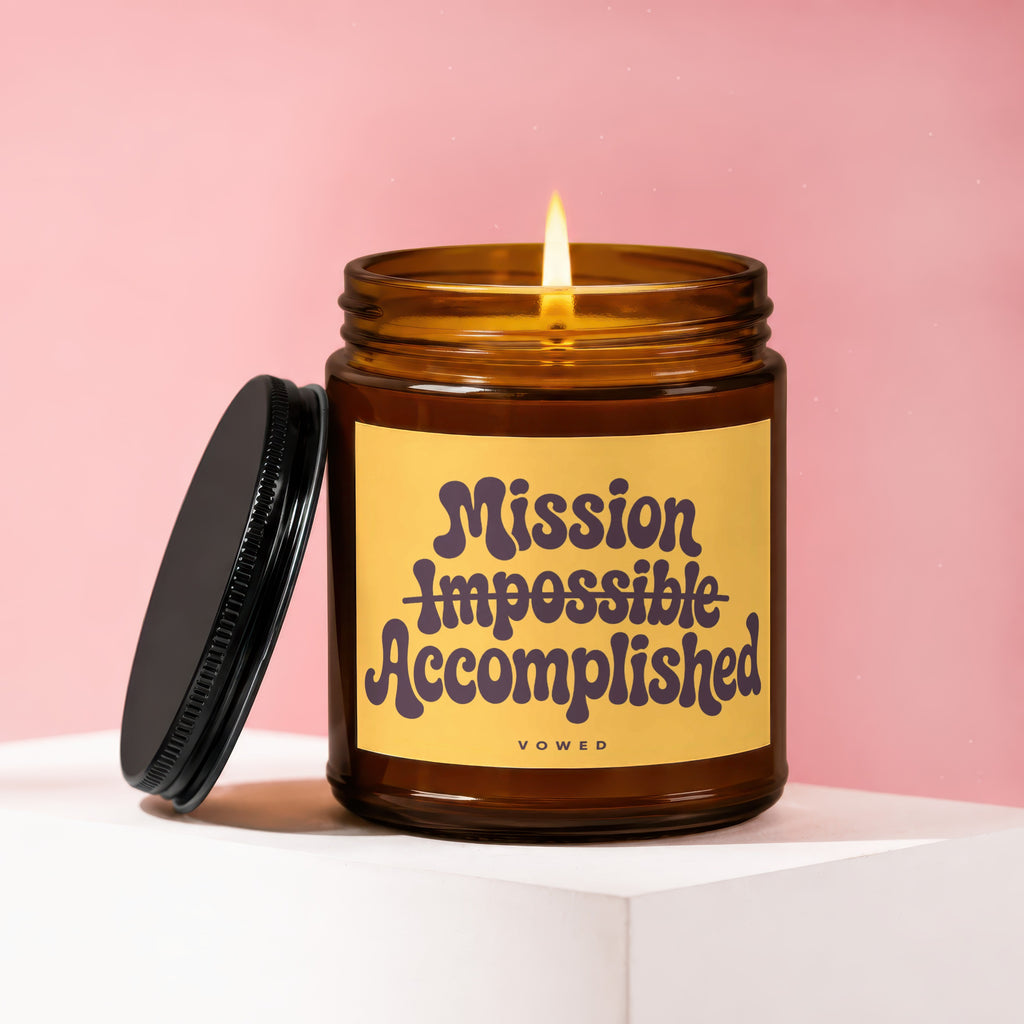 Amber glass candle with lit wick, yellow label reading "Mission Impossible Accomplished"