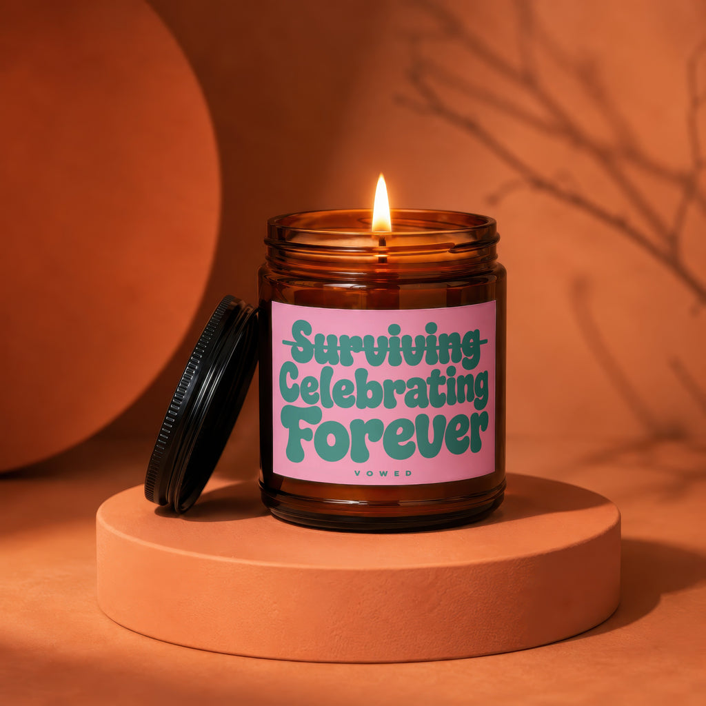 Amber candle with lit wick and pink label reading "Surviving Celebrating Forever Vowed."