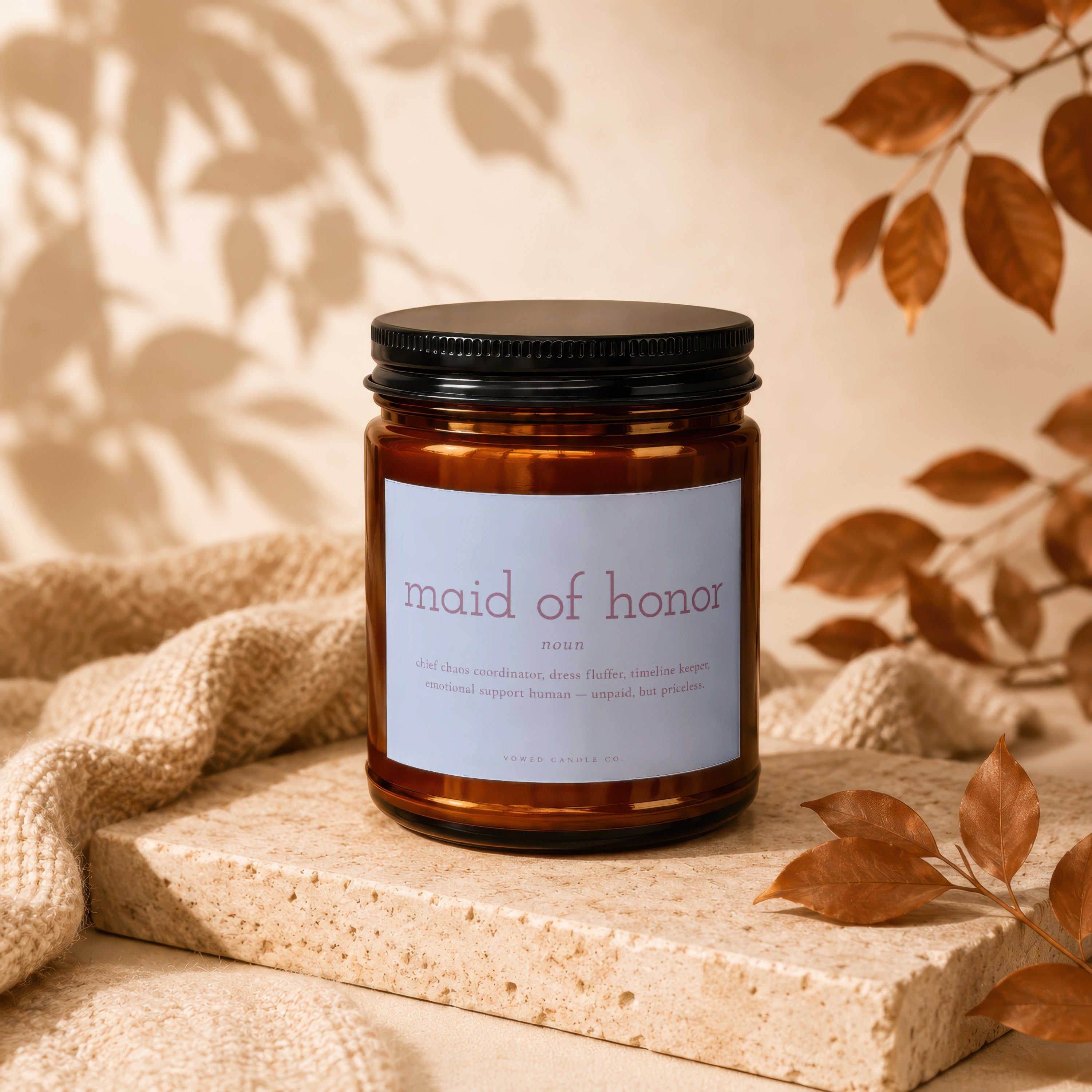 Amber jar candle labeled "maid of honor" with humorous definition, set on stone.