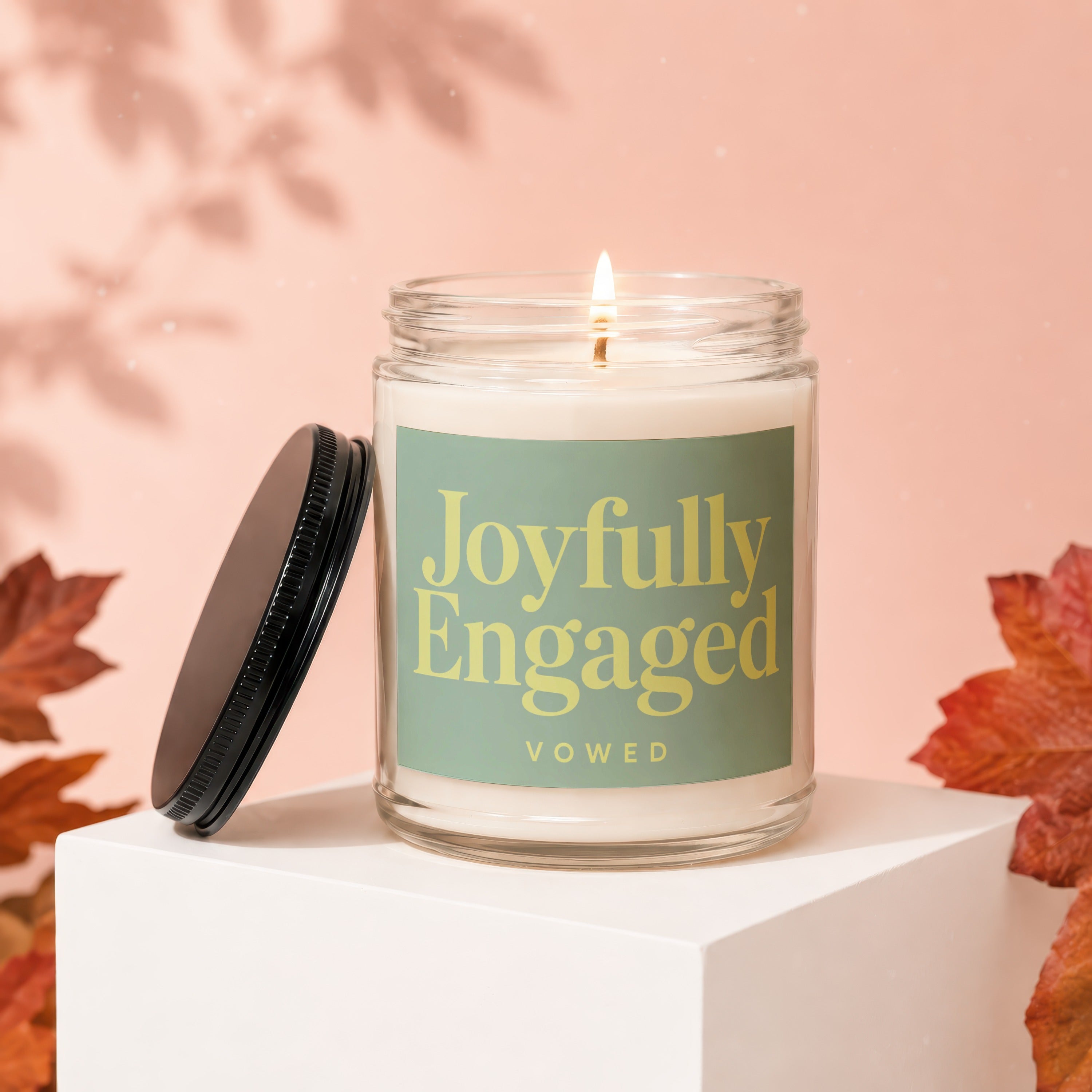 Candle with "Joyfully Engaged" text on a green label, lid off, on a white cube.