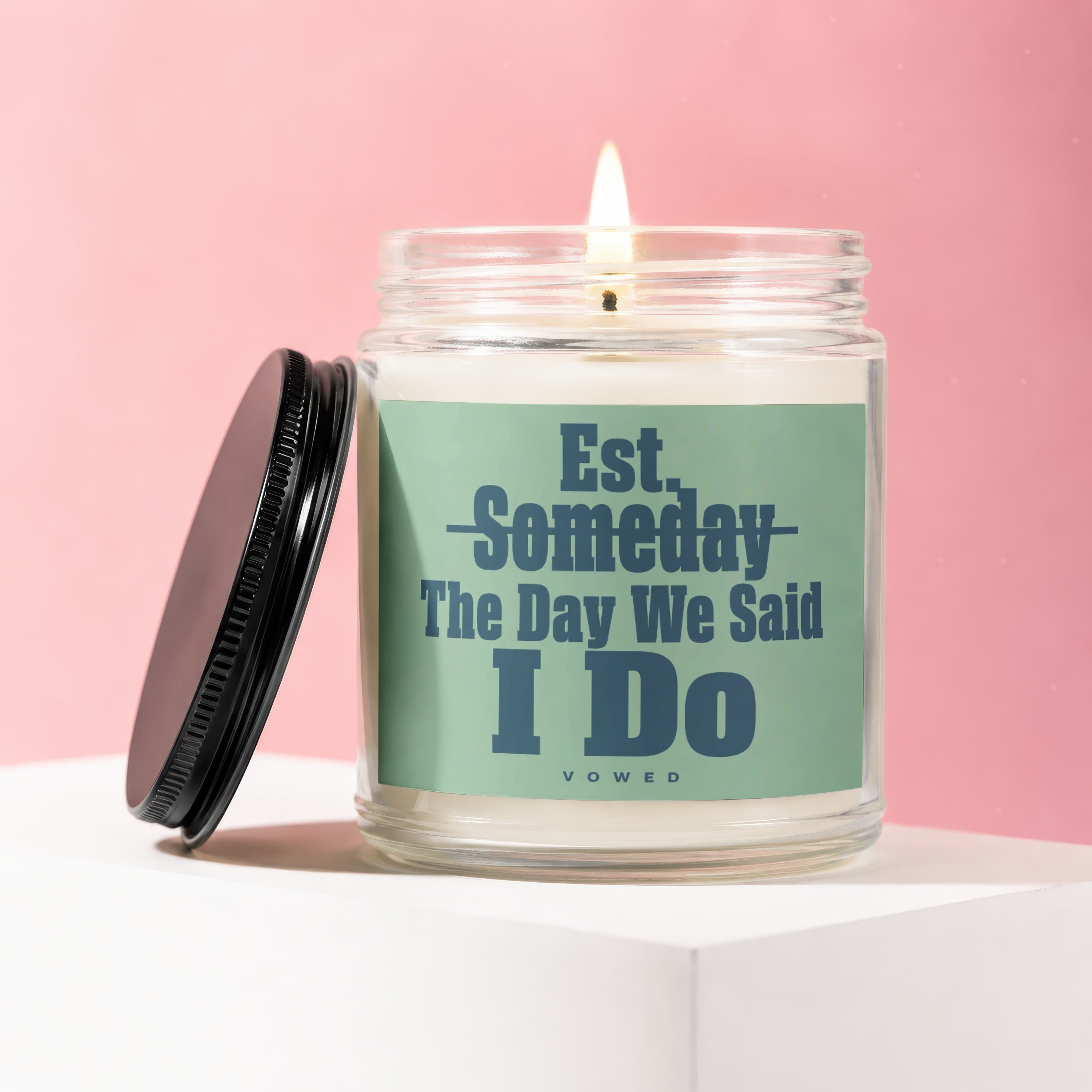 Glass candle with a lit wick and a teal label reading "Est. Someday The Day We Said I Do VOWED".