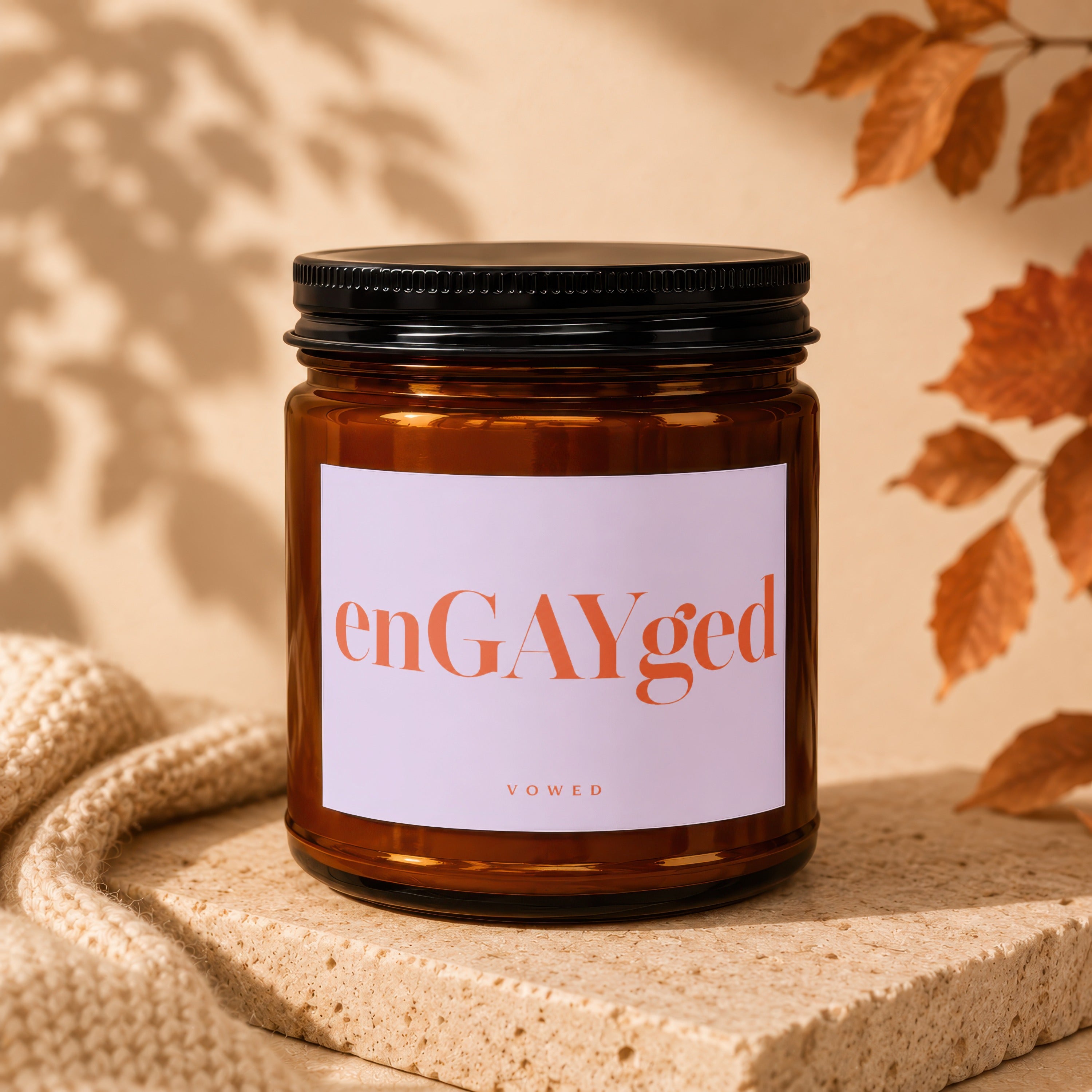 Amber jar candle with "enGAYged" label, beside knit fabric and autumn leaves.