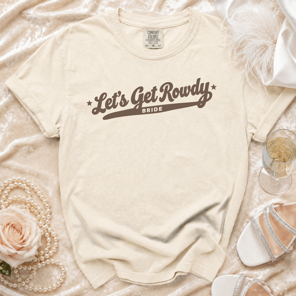 Cream-colored t-shirt with brown text reading Let's Get Rowdy Bride, laid flat among wedding accessories.