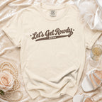 Cream-colored t-shirt with brown text reading Let's Get Rowdy Bride, laid flat among wedding accessories.