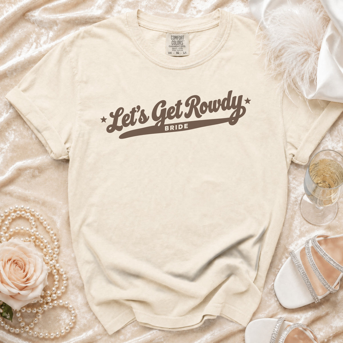 Cream-colored t-shirt with brown text reading Let's Get Rowdy Bride, laid flat among wedding accessories.