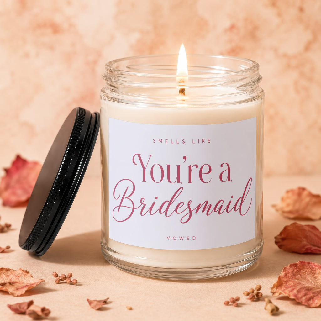 Candle with lit wick, glass jar, and label reading "Smells Like You're a Bridesmaid Vowed".