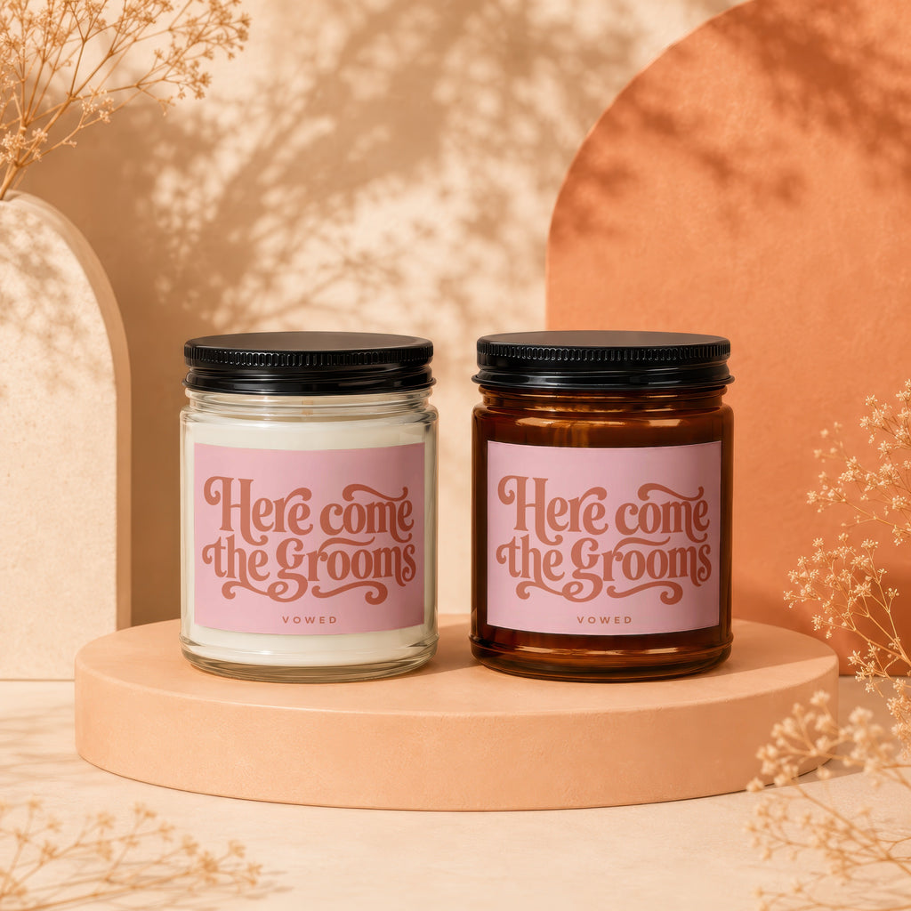 Two candles with pink labels reading "Here come the grooms" displayed on risers.