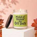 Glass candle with "Maid of Honor on Off Duty" text, lid off, lit wick.