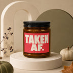 Amber candle with black lid, red label with "TAKEN AF. VOWED" text.