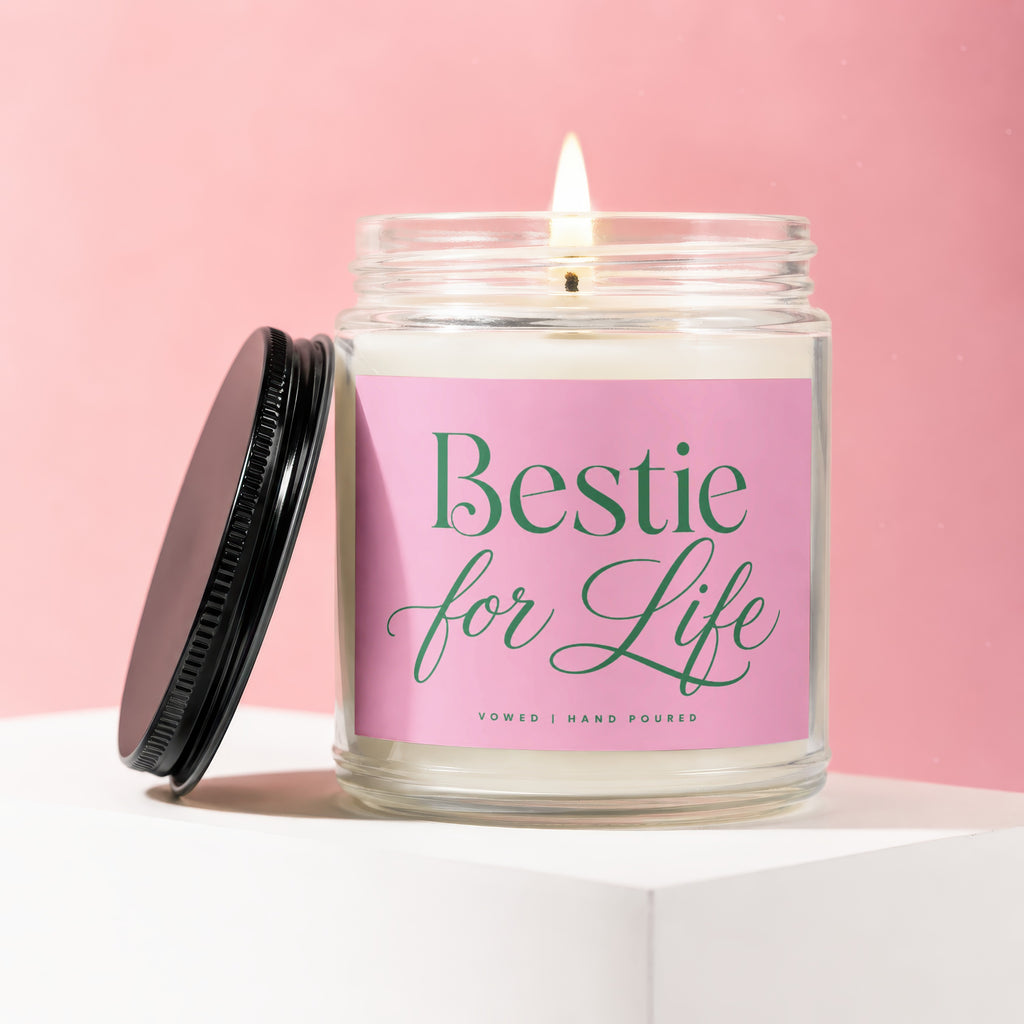 Glass candle with "Bestie for Life" label and lit wick on a pink background.