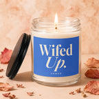 Glass candle burning with "Wifed Up. VOWED" on blue label, lid to the left.