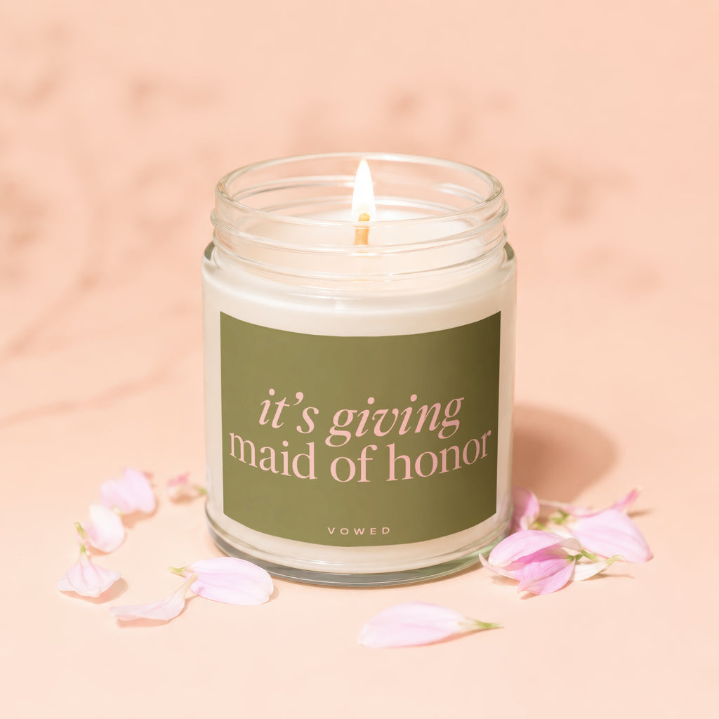 Candle with a burning wick and green label that reads "it's giving maid of honor".