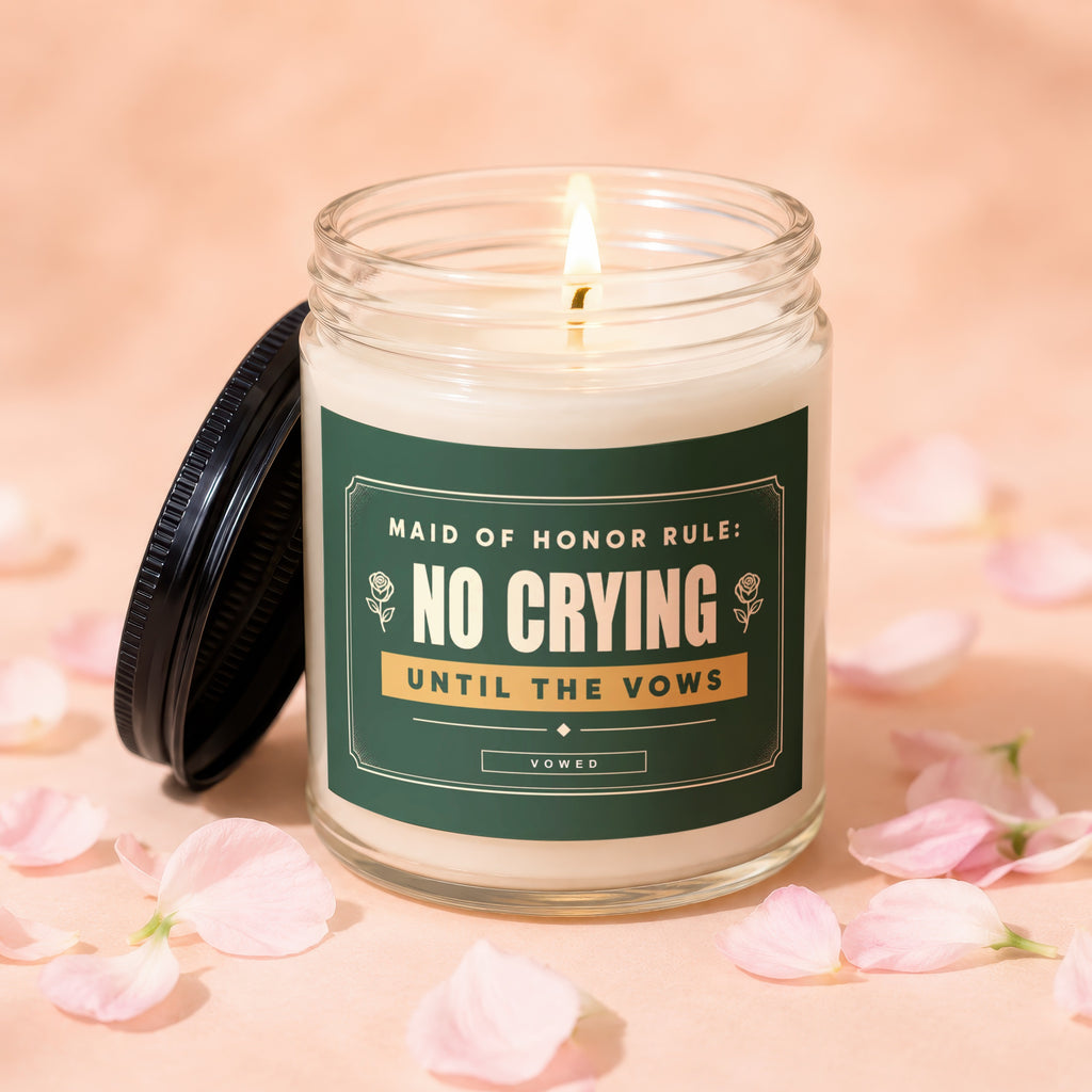 Glass jar candle with green label reading "NO CRYING UNTIL THE VOWS" surrounded by pink petals.