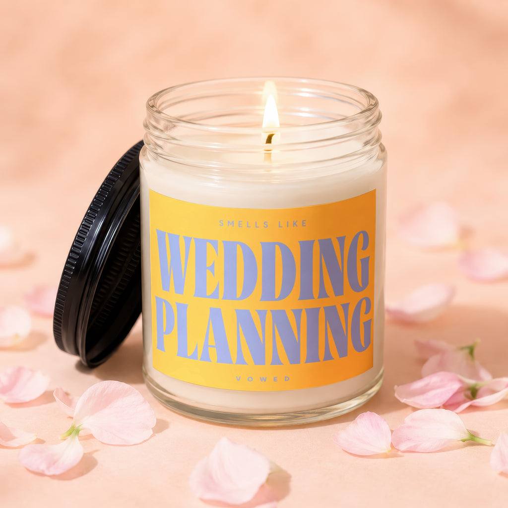 Lit candle in glass jar with yellow label reading "Sells Like Wedding Planning Vowed".