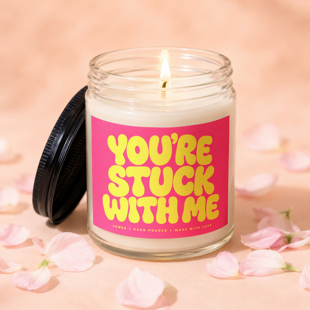Glass candle with burning wick and pink label reading "YOU'RE STUCK WITH ME".
