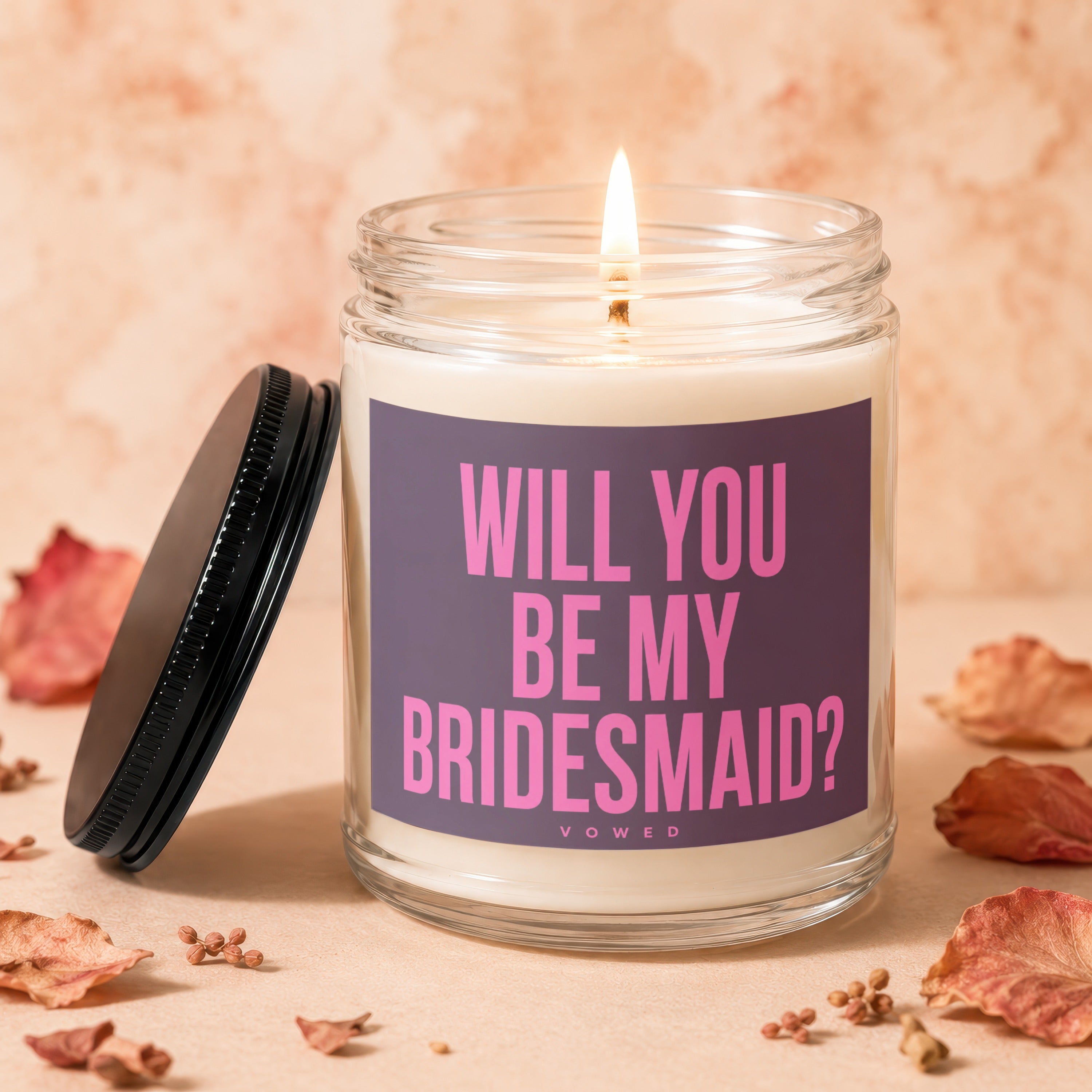 Candle with lit wick and "Will You Be My Bridesmaid?" text on label.
