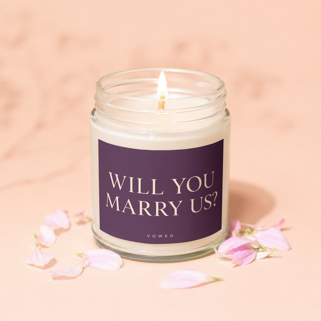 Lit candle in jar with "Will You Marry Us?" text and pink petals.