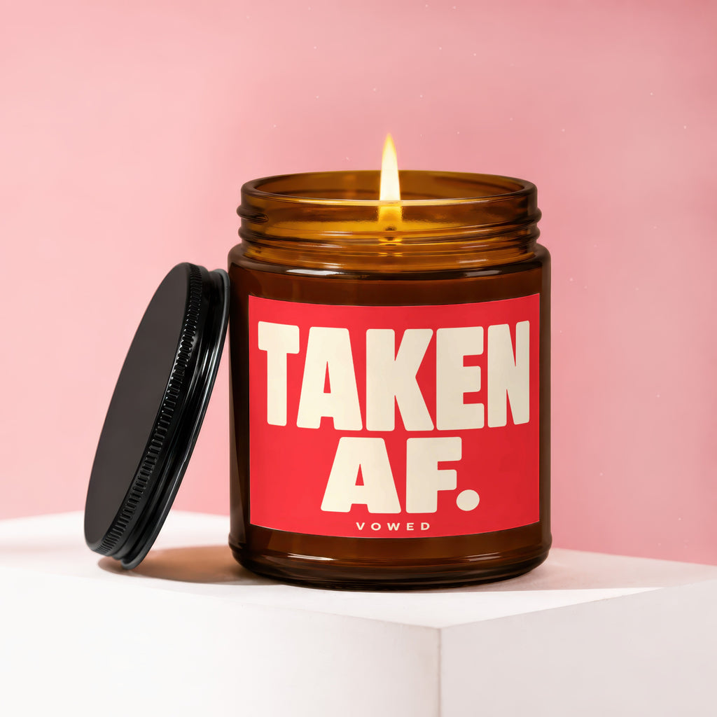 Amber jar candle with lit wick and red label reading "TAKEN AF. VOWED."