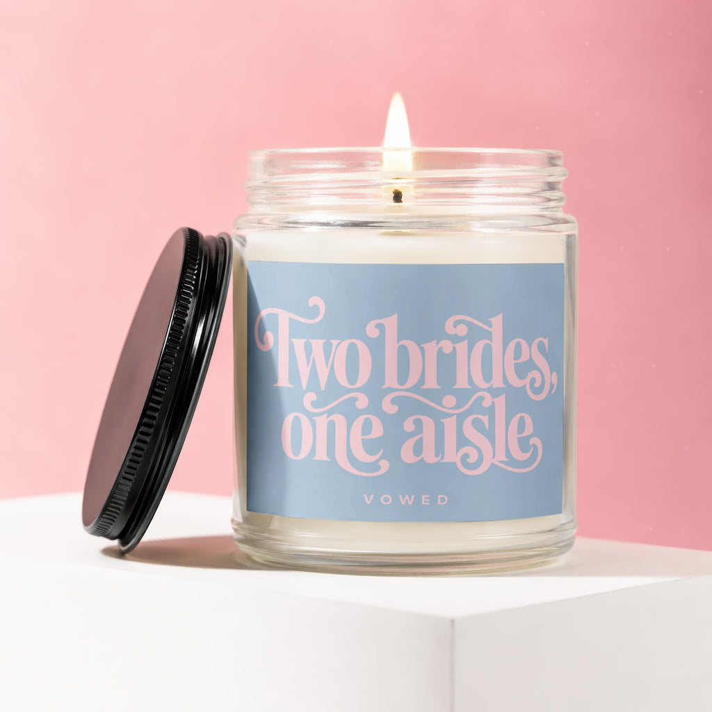 Lit candle in glass jar with blue label reading "Two brides, one aisle."