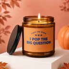 Amber jar candle with "LIGHT ME WHEN I POP THE BIG QUESTION" text and a lit wick.