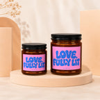 Two amber glass candles with pink labels reading "LOVE, FULLY LIT"