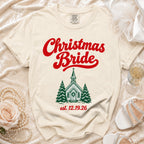 Cream t-shirt featuring red script text reading "Christmas Bride", a green church graphic, and the date "est. 12.19.26".