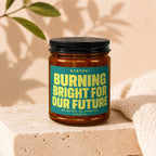 Amber jar with black lid, teal label with yellow text: "STATUS: BURNING BRIGHT FOR OUR FUTURE VOWED".