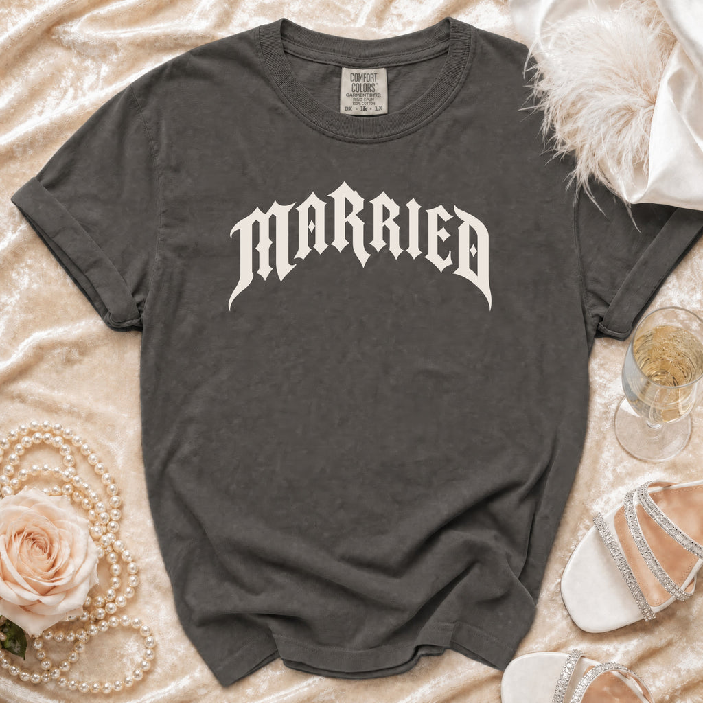 A dark gray t-shirt with the word MARRIED in white gothic lettering, laid on a cream velvet background with wedding accessories.