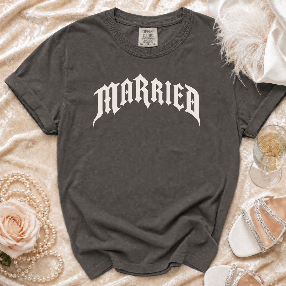 A dark gray t-shirt with the word MARRIED in white gothic lettering, laid on a cream velvet background with wedding accessories.