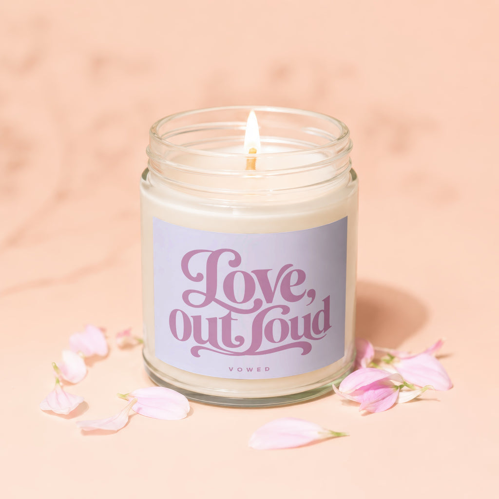 Glass jar candle with a lit wick and a "Love, out loud" label surrounded by flower petals.
